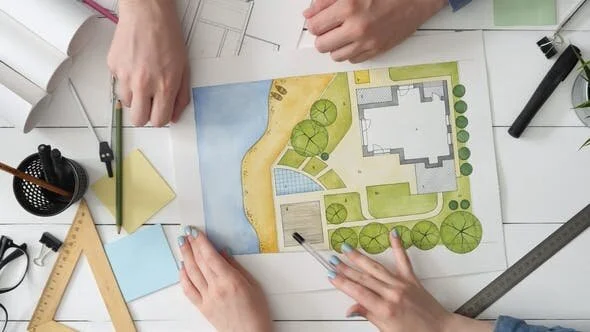 Top-down view of hands working on a colorful hand-drawn landscape architecture plan for a residential property with a pool and garden.