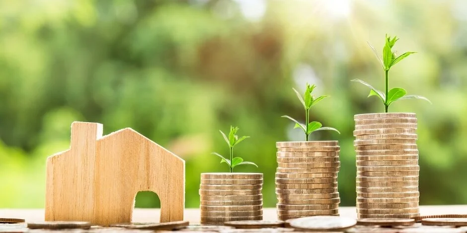 A wooden house model next to rising stacks of coins with growing green sprouts against a blurred nature background.