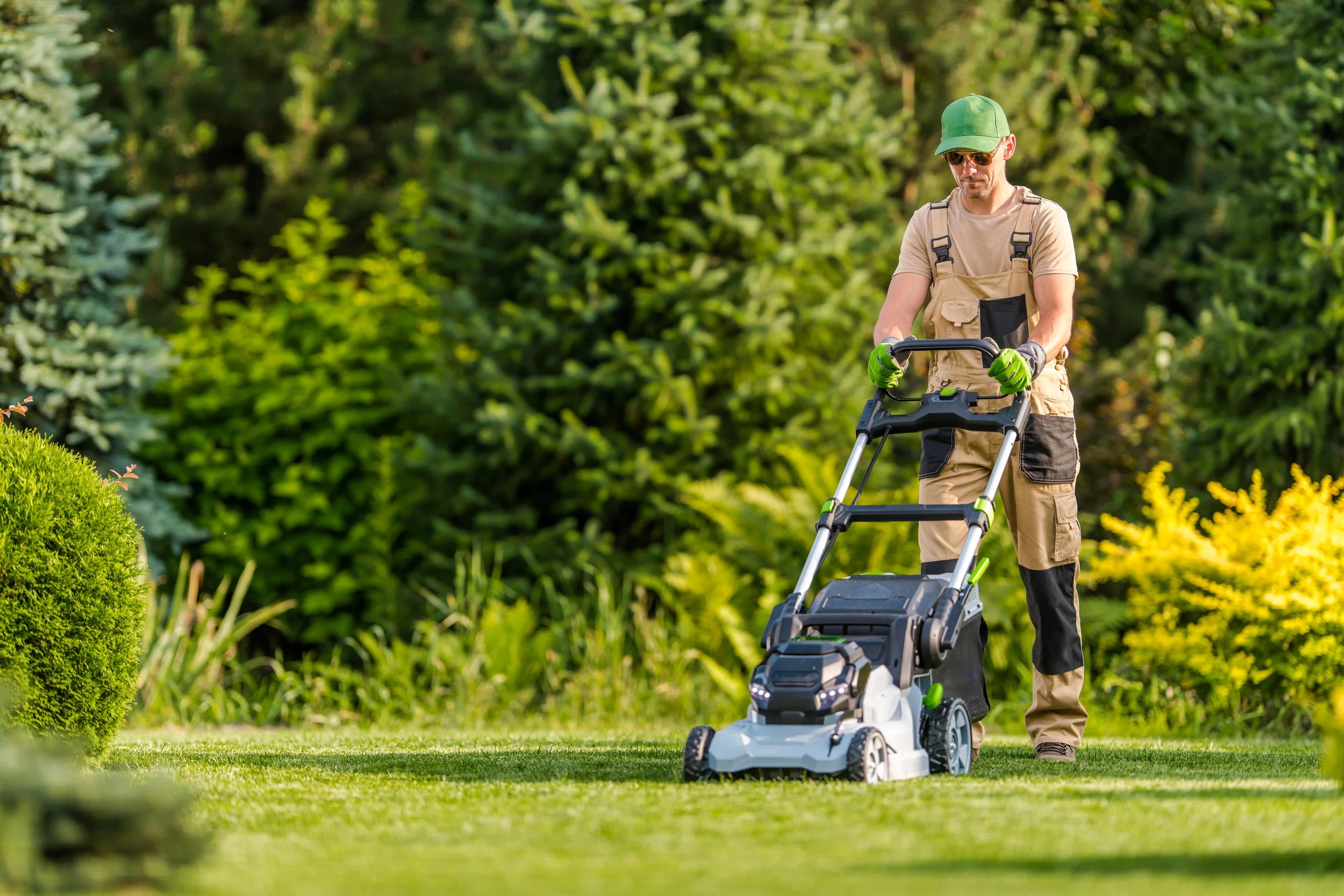 A professional landscaper wearing a green cap and tan overalls operating a modern push mower on a bright, sunny lawn.