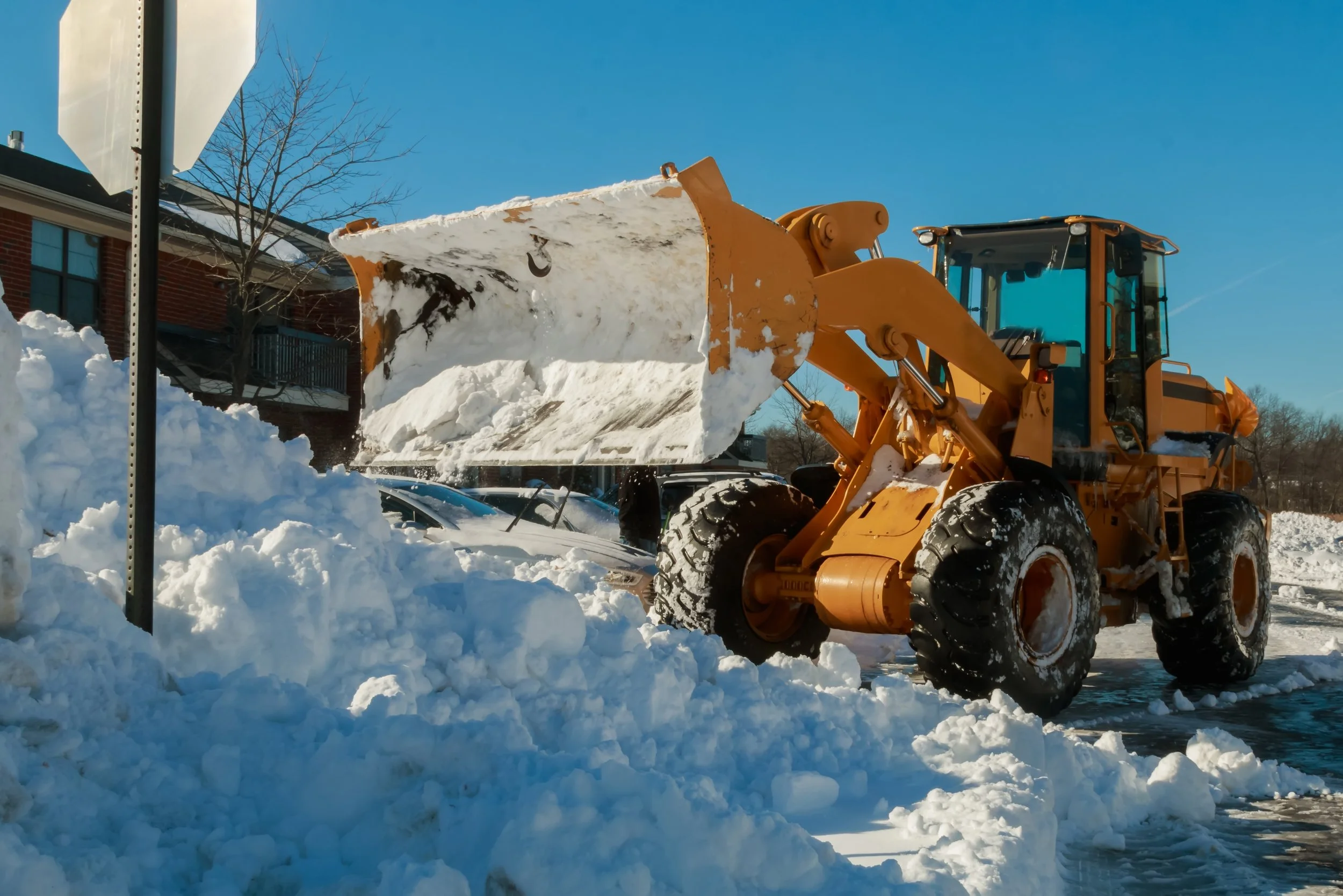 A large yellow front-end loader lifting a bucket full of snow while clearing a parking lot or residential street on a sunny day.