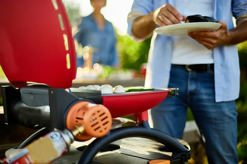A man grilling food on a portable red gas grill outdoors during a backyard barbecue.