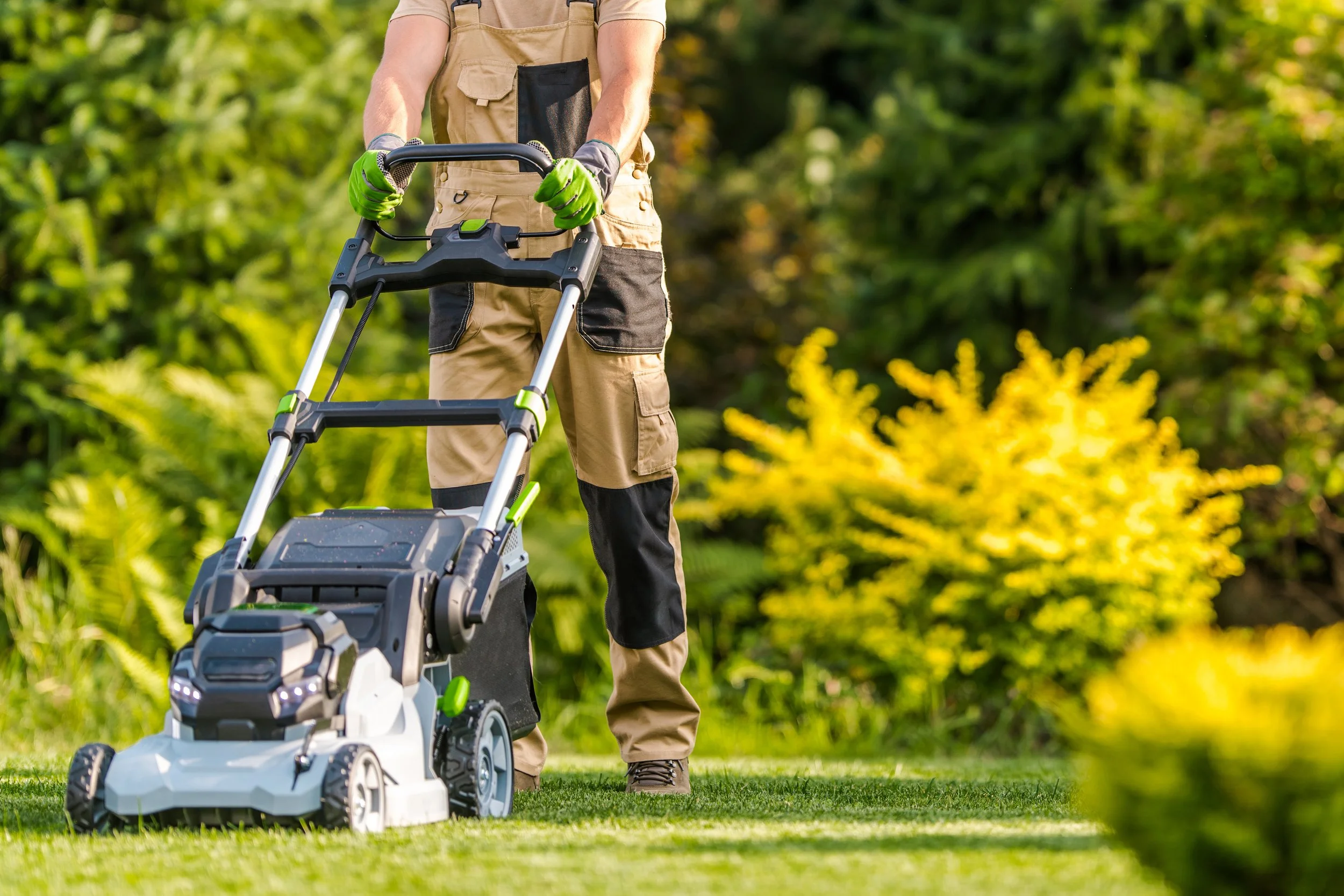 A professional landscaper in tan and black work overalls and green gloves operates a modern white and black lawnmower on a lush, well-maintained green lawn.