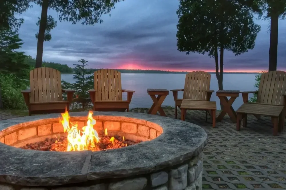 Stone fire pit with glowing flames and Adirondack chairs on a lakeside patio at sunset