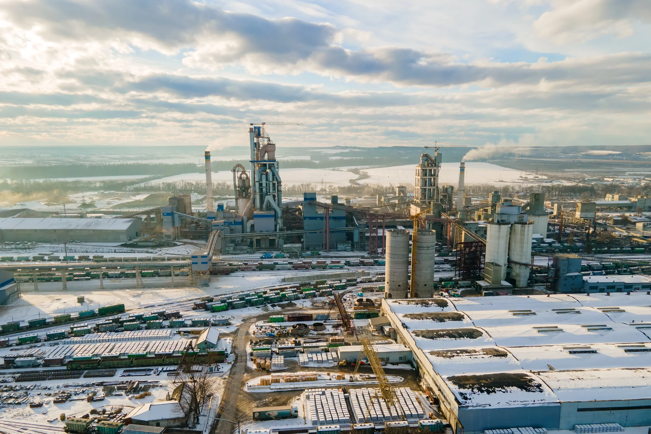 Aerial view of a large industrial factory complex with smokestacks and silos in a snowy landscape under a cloudy sky.