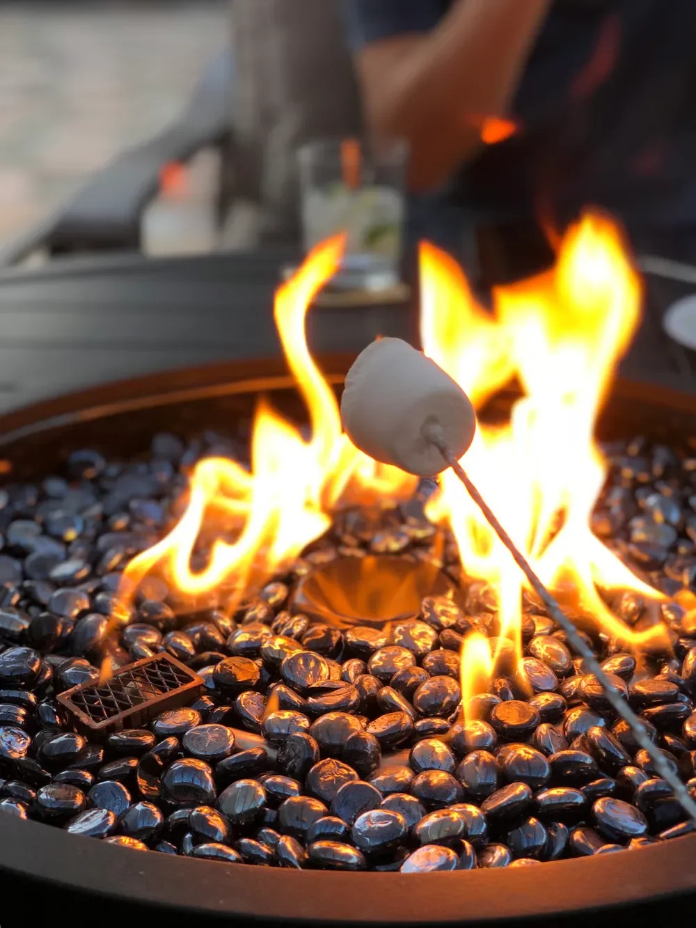 Marshmallow roasting on a skewer over bright flames in a gas fire pit filled with black fire glass rocks.
