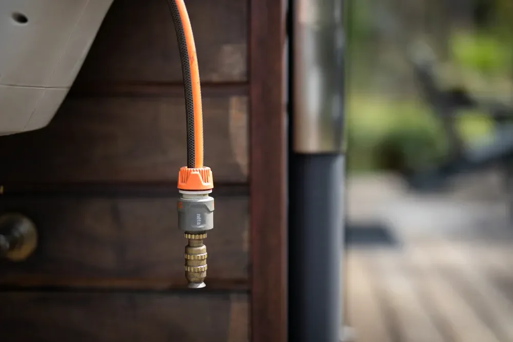 An orange and black garden hose with a brass quick-connect adapter hanging outdoors against a wooden wall.