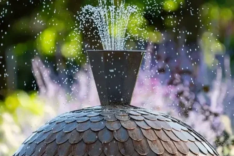 Dark stone garden water fountain spraying water from the top, with blurred purple flowers in the background.