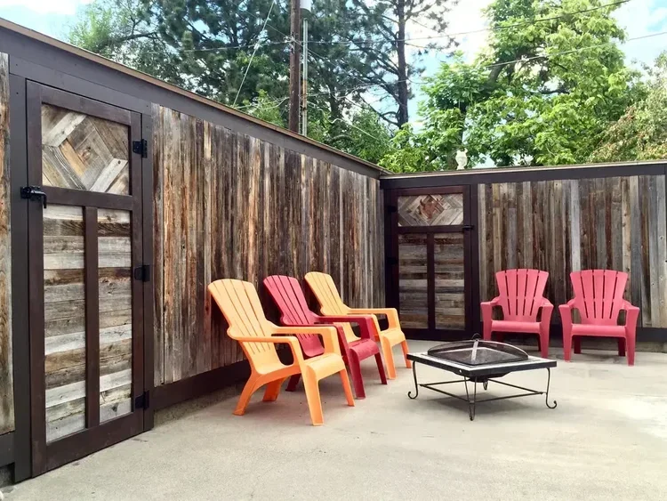 Colorful chairs and a metal fire pit set on a concrete patio enclosed by a rustic wooden privacy fence with gate.