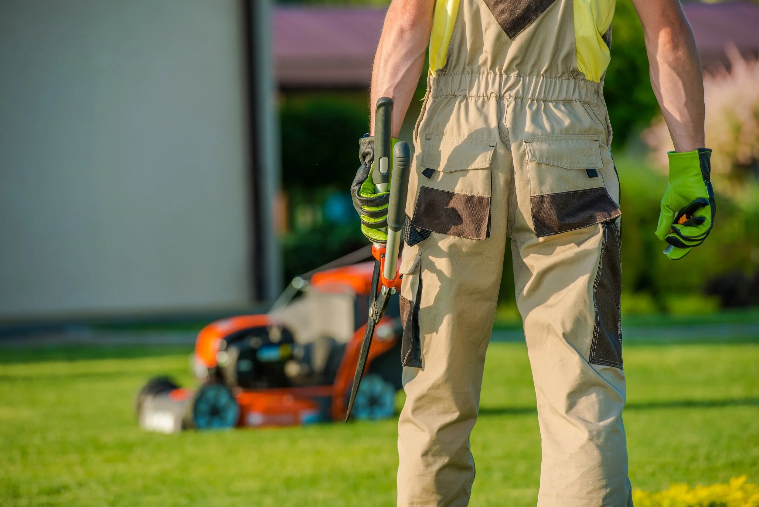 A landscaper in tan work overalls and green gloves stands on a lawn with an orange lawn mower in the background.
