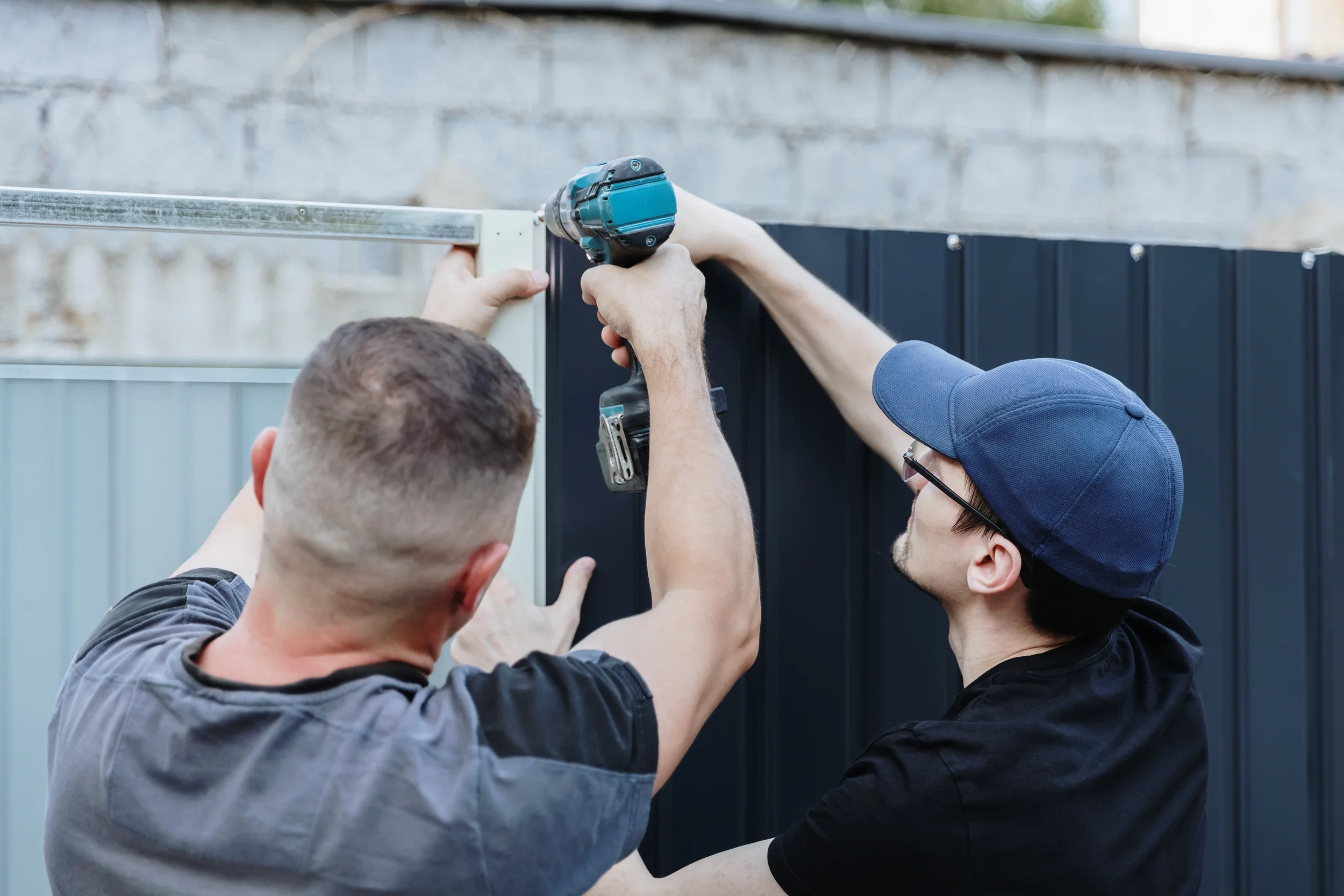 Two professional contractors using a power drill to install a modern dark metal privacy fence on a residential property.