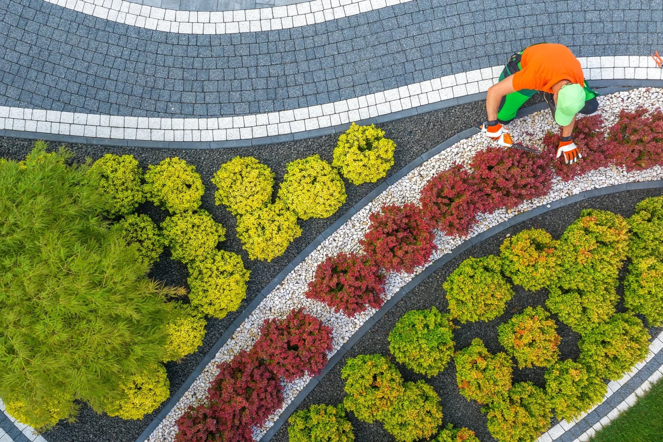 Aerial view of a landscaper maintaining a curved garden bed with colorful shrubs and decorative stone pavers.