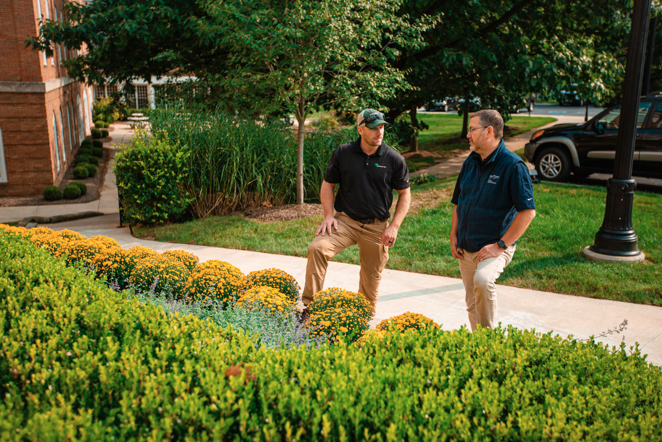 Two men stand on a paved path in a lush garden, discussing landscaping near a row of yellow flowers and green hedges.