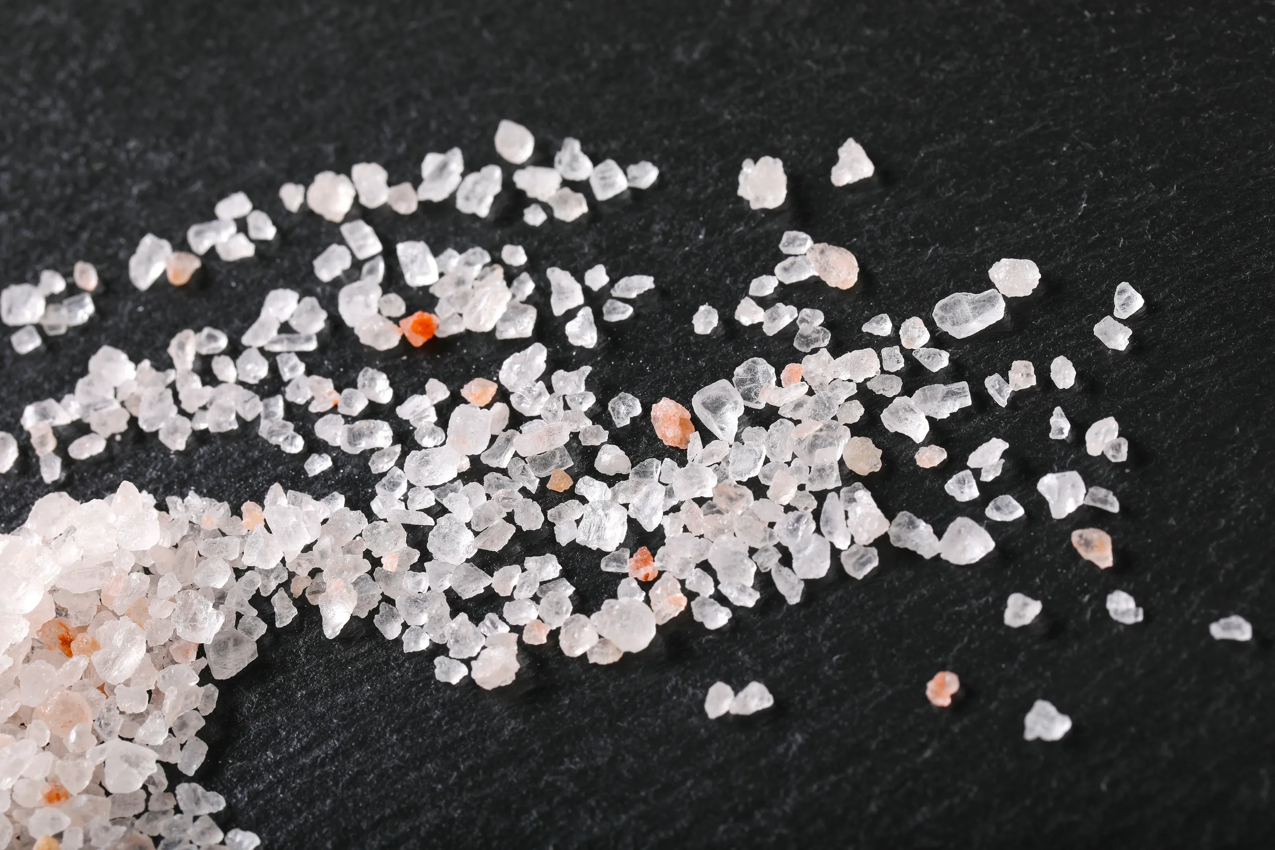 Coarse white and pink rock salt crystals scattered on a dark asphalt surface for winter ice melting.