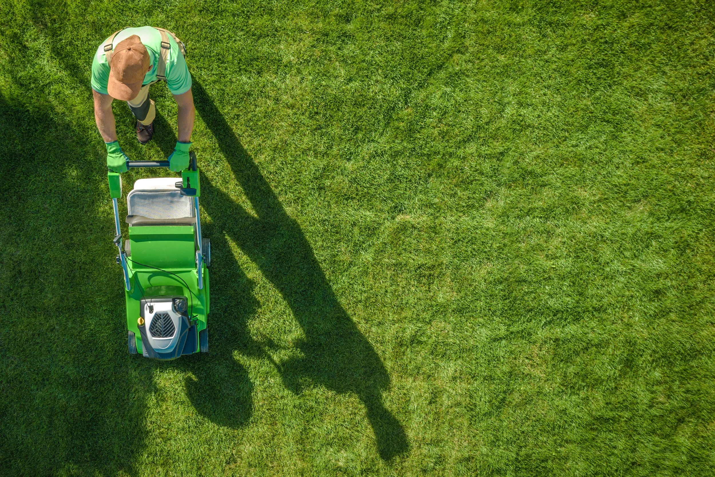 Top-down aerial view of a professional landscaper in a green shirt and brown cap mowing a lush, vibrant green lawn.