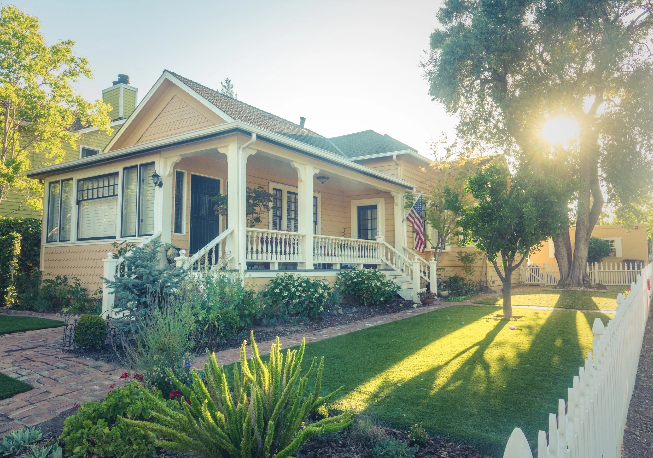 A sunlit, pale yellow craftsman house with a white wraparound porch, a small American flag, and a white picket fence under a glowing afternoon sun.
