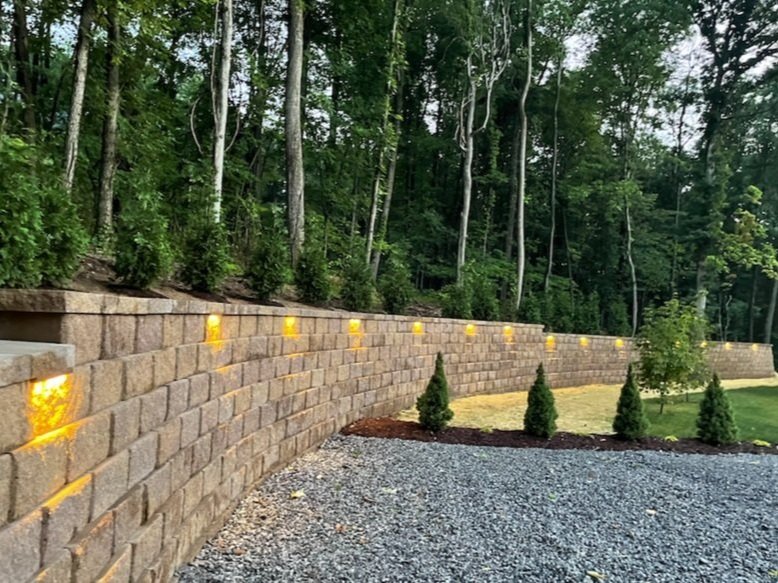 Long stone retaining wall featuring integrated LED cap lights at dusk, bordering a gravel path and wooded area.
