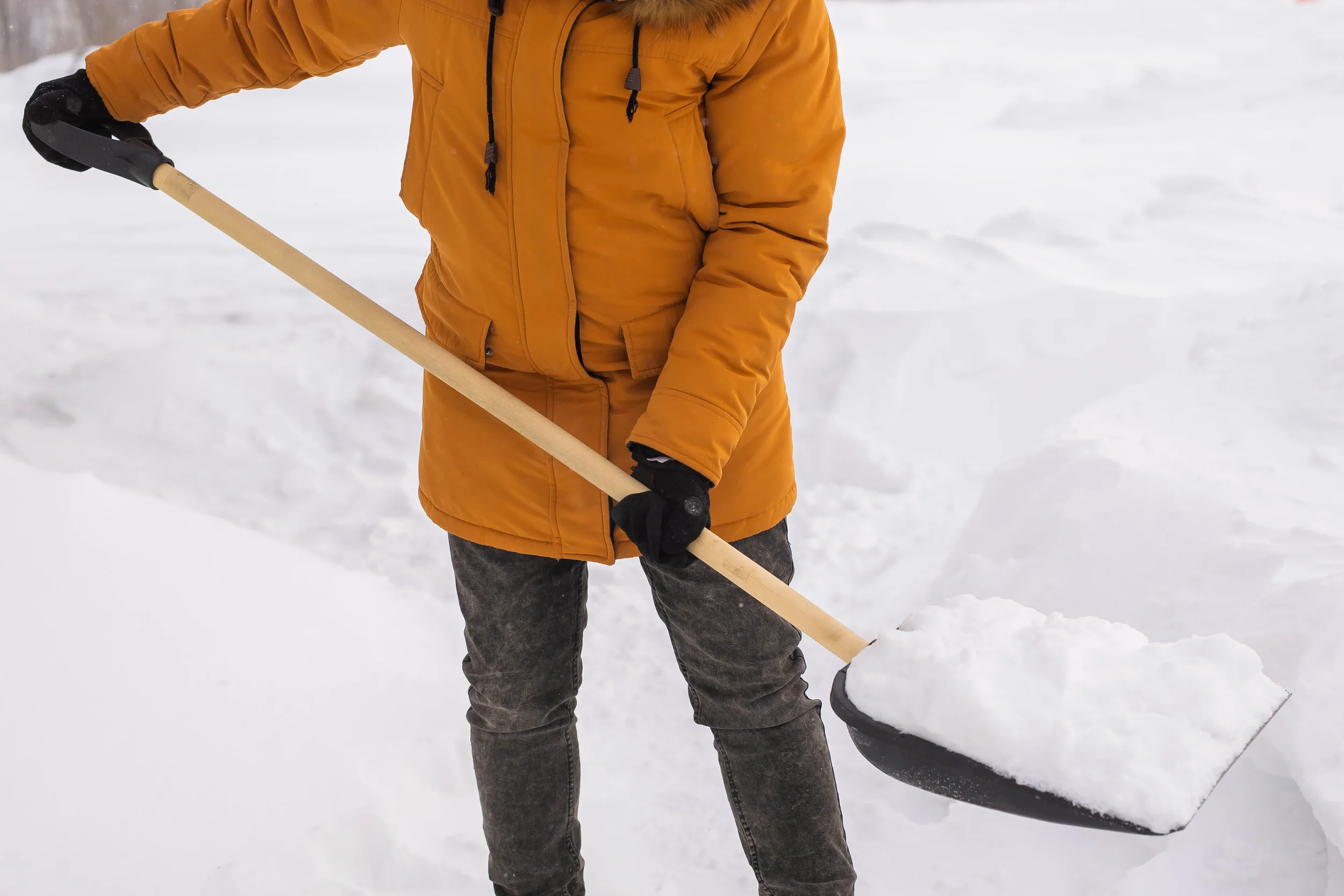 Person in an orange winter parka shoveling heavy white snow from a driveway during a cold winter day.