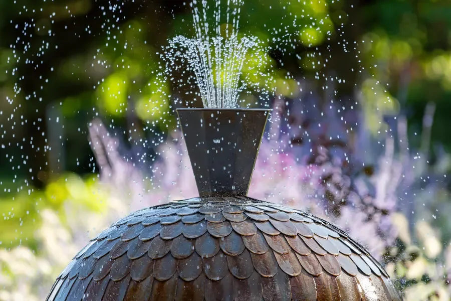 Scalloped bronze garden fountain nozzle spraying water outdoors with blurred flowers.