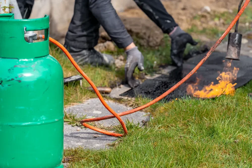 A worker uses a propane torch connected to a green gas tank to burn weeds and clear a stone walkway path.
