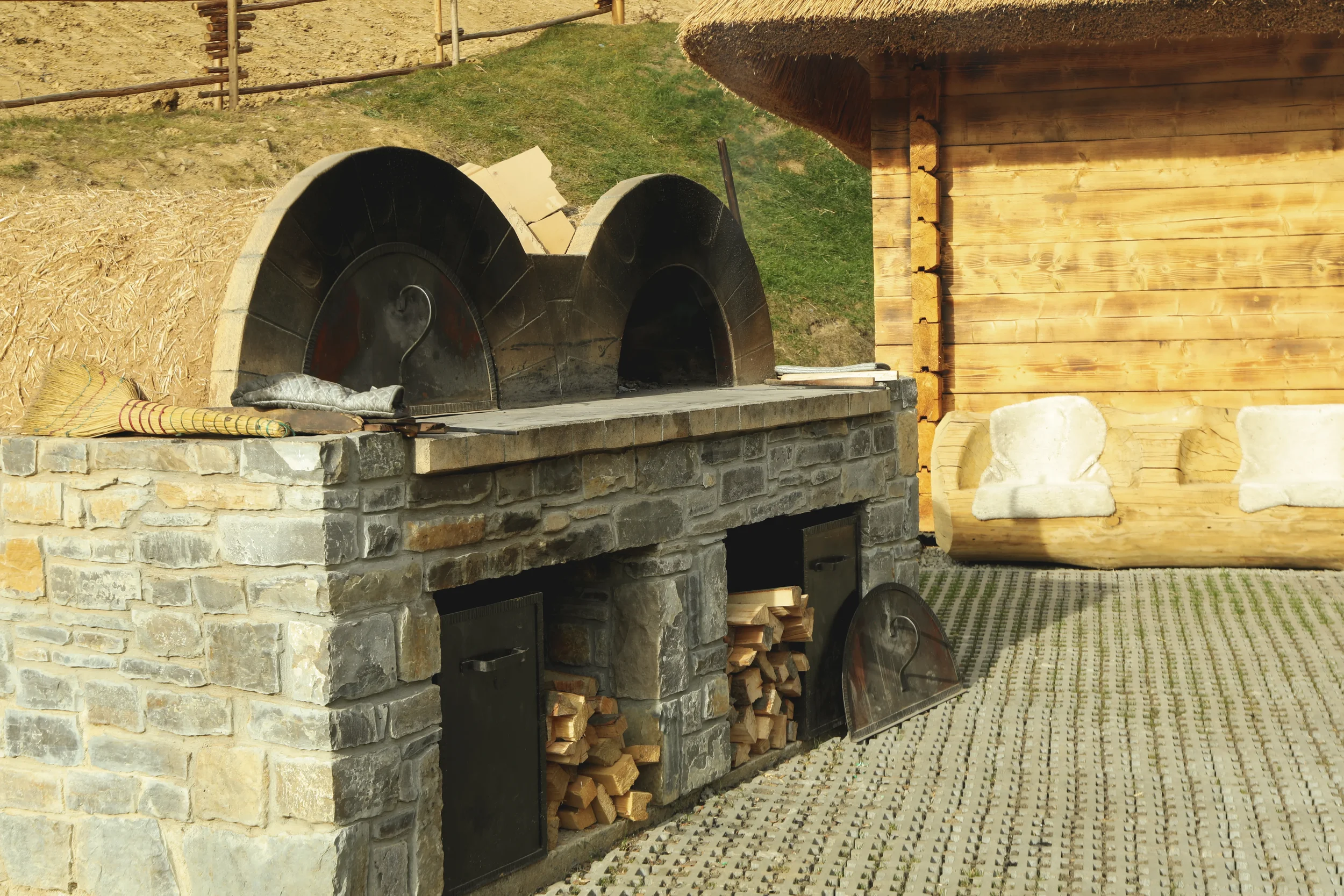 Rustic stone outdoor oven with two arched ovens and firewood storage beside a log cabin