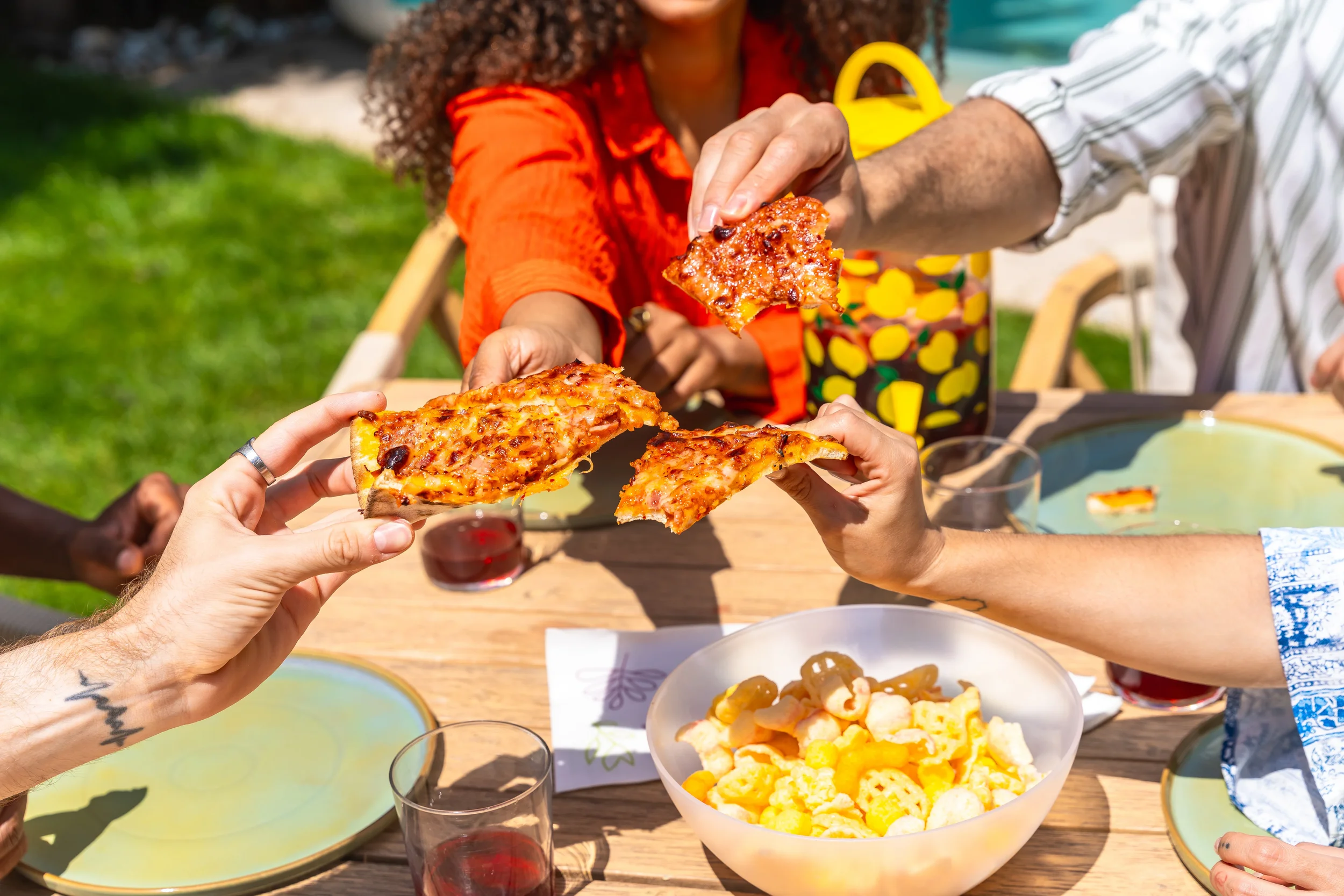 Friends sharing slices of homemade pizza with red wine at a sunny outdoor backyard table