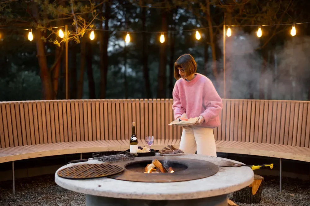 Cozy backyard patio with a concrete fire pit, large curved wooden bench, and warm overhead string lights at night.