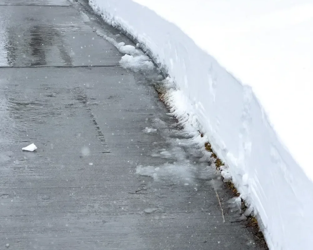 A clear, wet concrete sidewalk bordered by a high, neatly shoveled bank of white snow during winter.