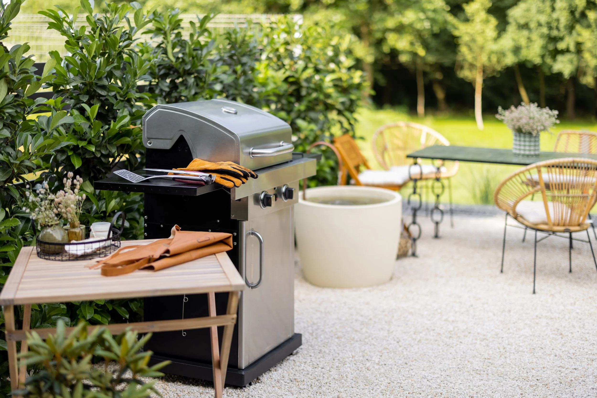 Stainless steel gas grill on a gravel patio with a wooden side table, outdoor furniture, and green privacy hedge.