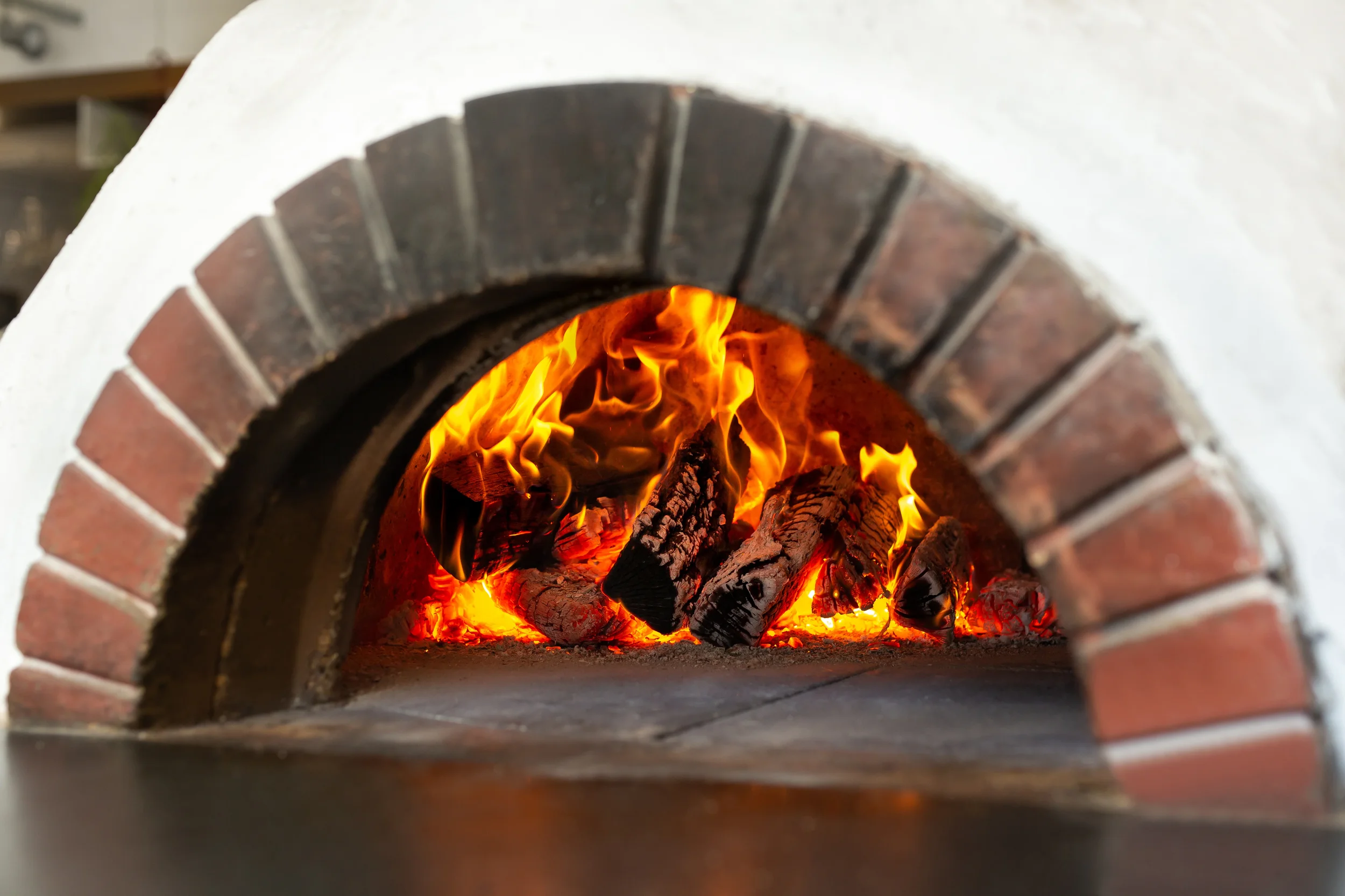 Roaring fire with burning logs inside a white wood-fired pizza oven with a red brick arch