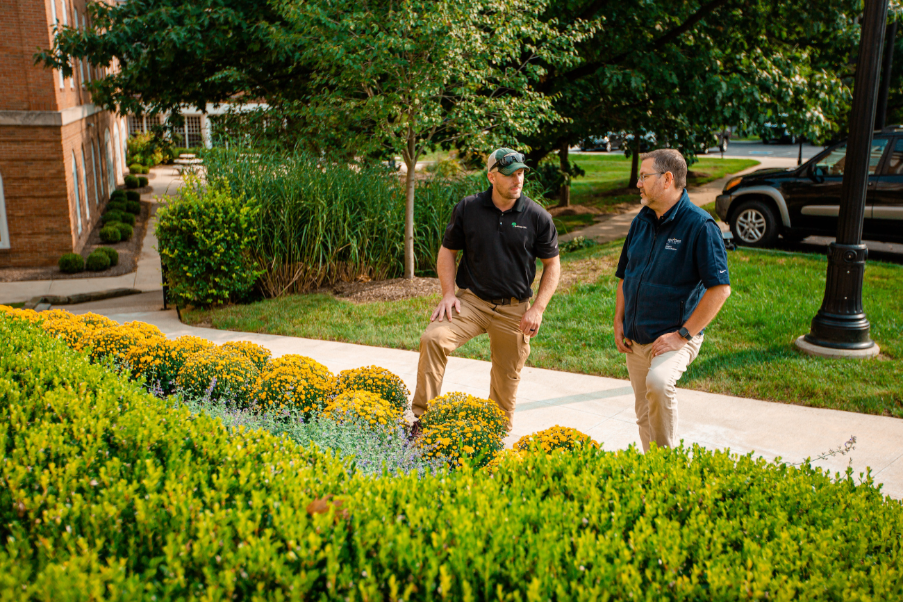 Two landscaping professionals discussing property maintenance near a commercial building with yellow flowers.