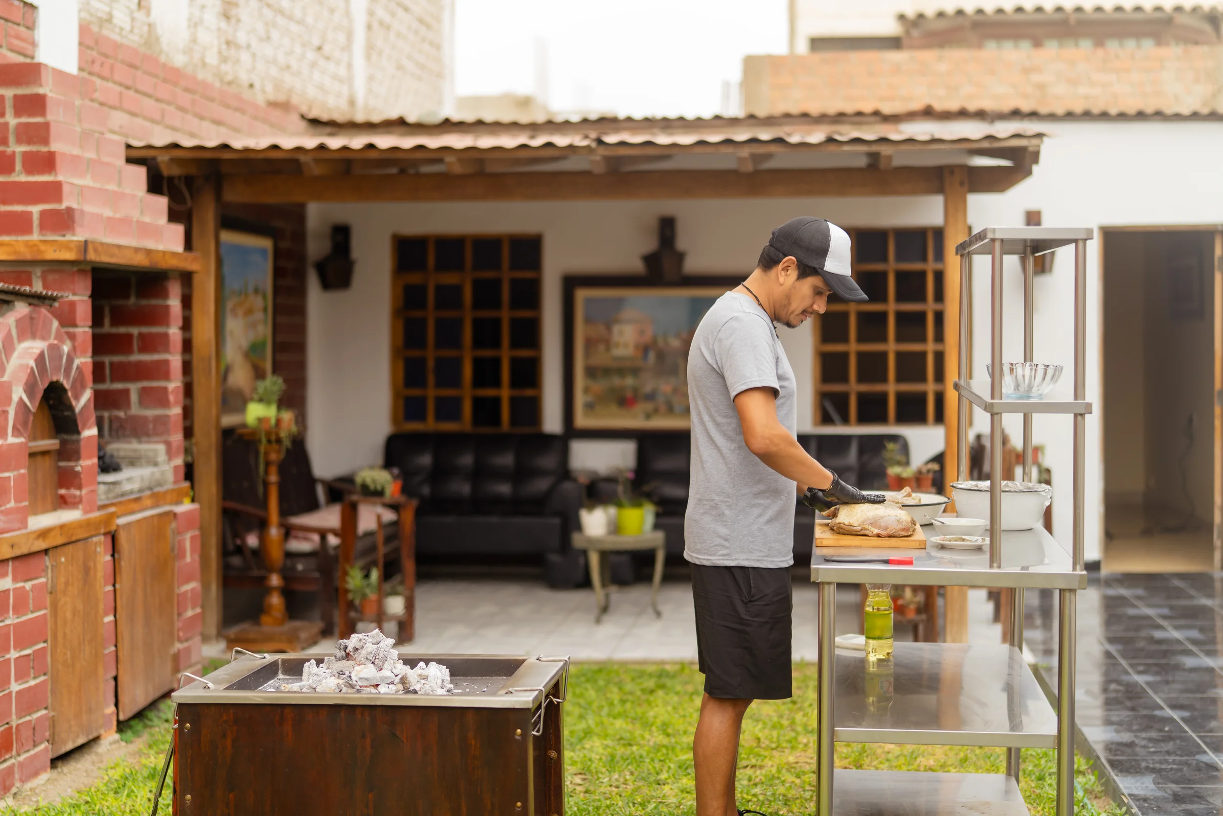 Man preparing food at a stainless steel prep cart beside a red brick pizza oven in a backyard