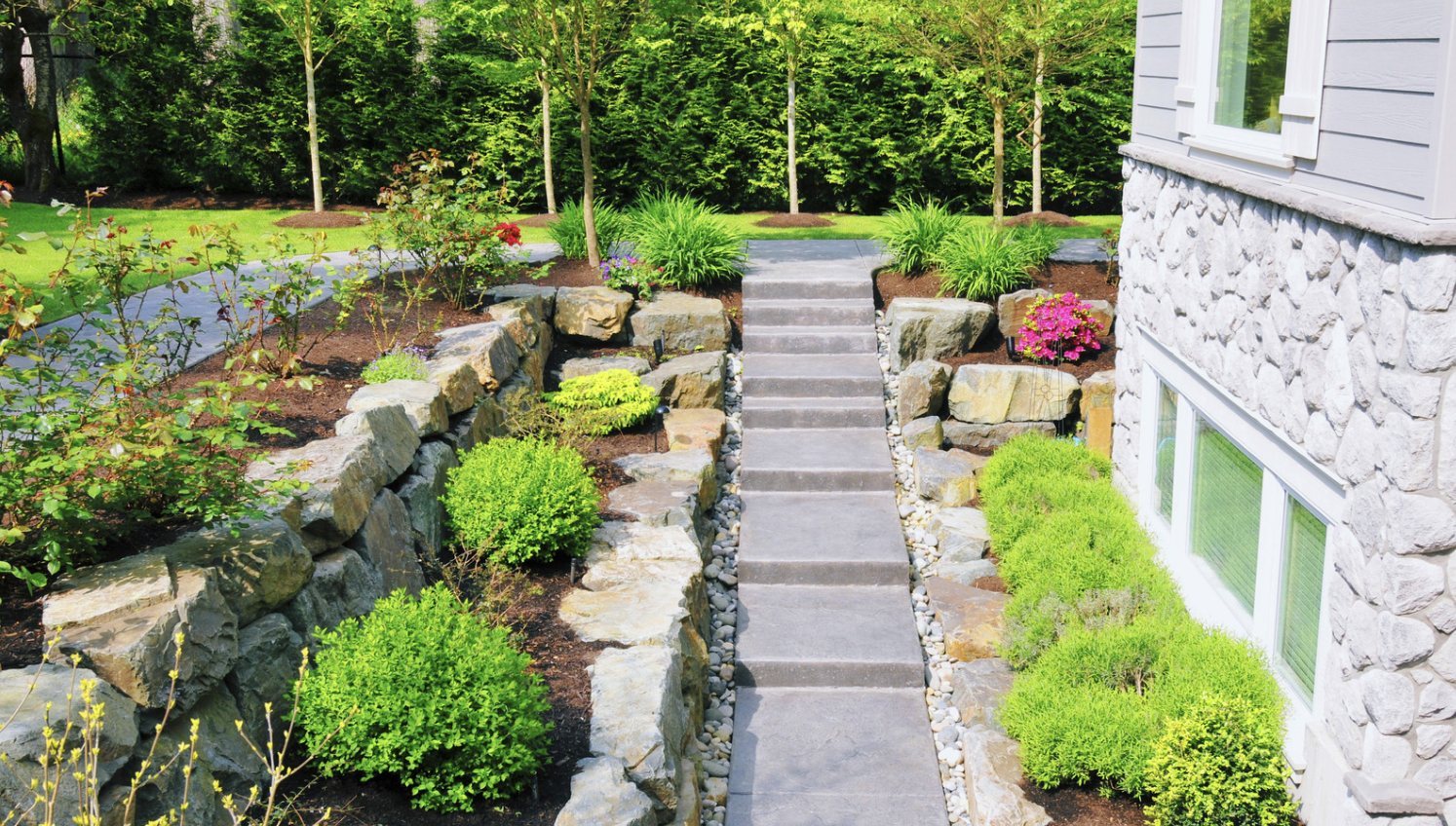 A series of gray stone steps leads up a sloped garden featuring a stone retaining wall, vibrant green shrubs, and pink flowers next to a light-colored house.
