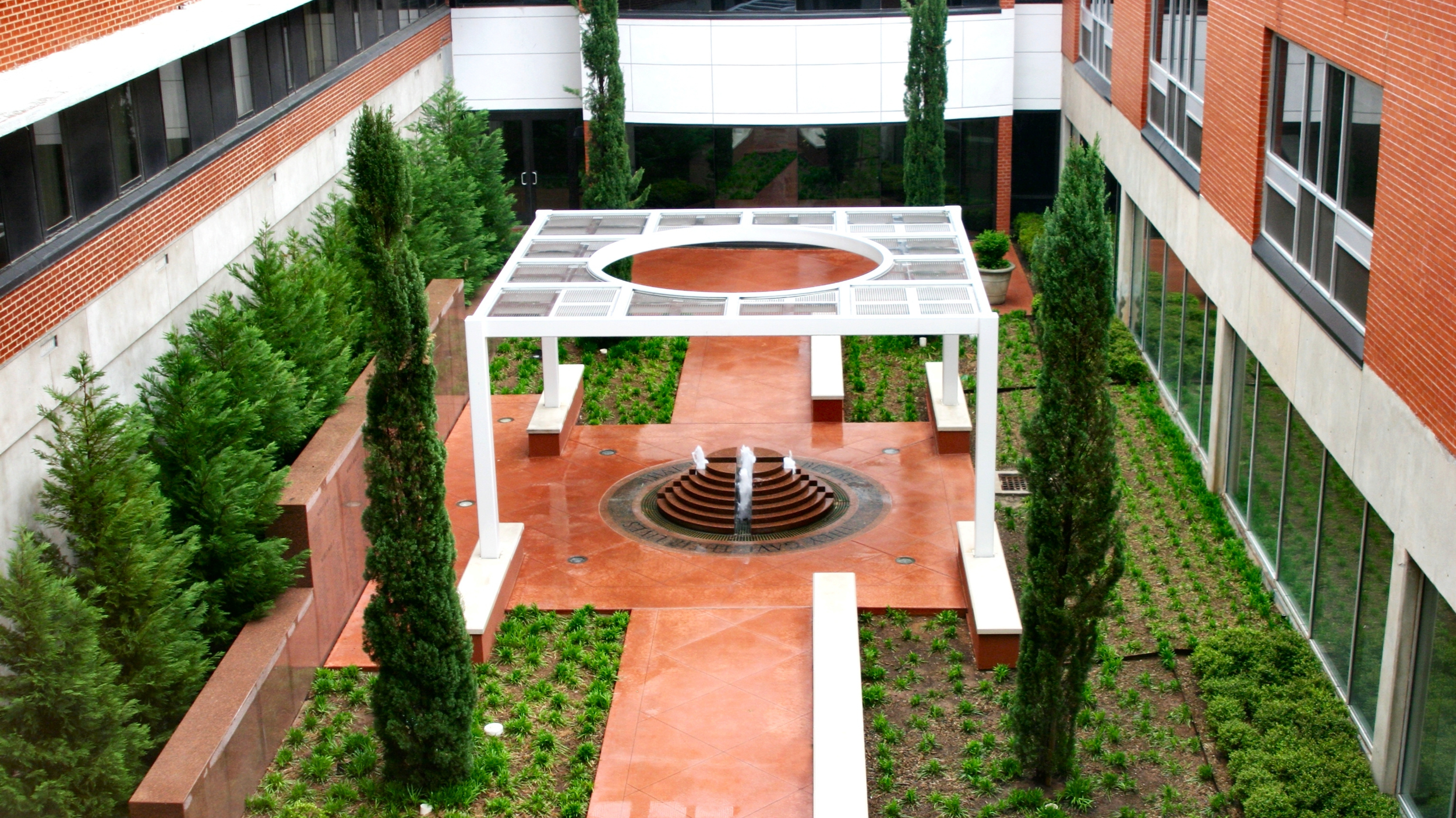An aerial view of a modern courtyard with a white pergola, circular fountain, terracotta paths, and tall evergreen trees.