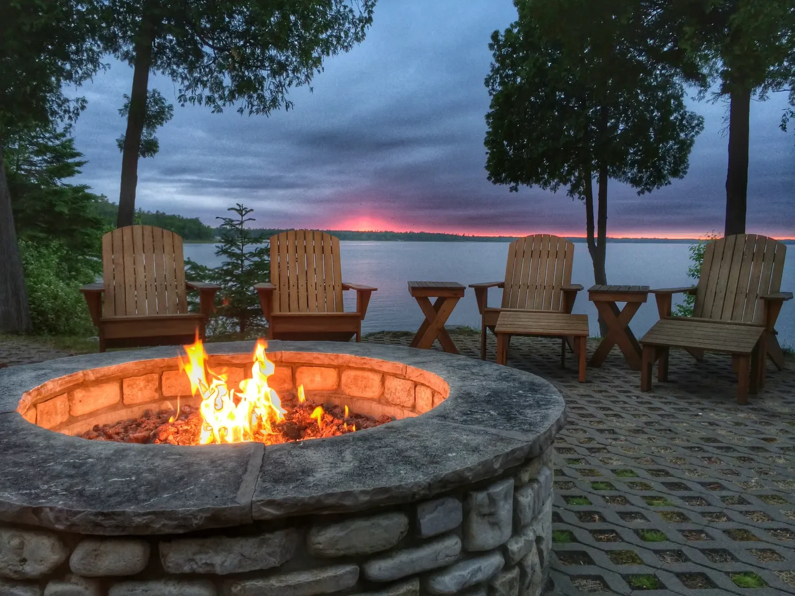 Stone fire pit burning on a paver patio with wooden chairs overlooking a serene lake at sunset.