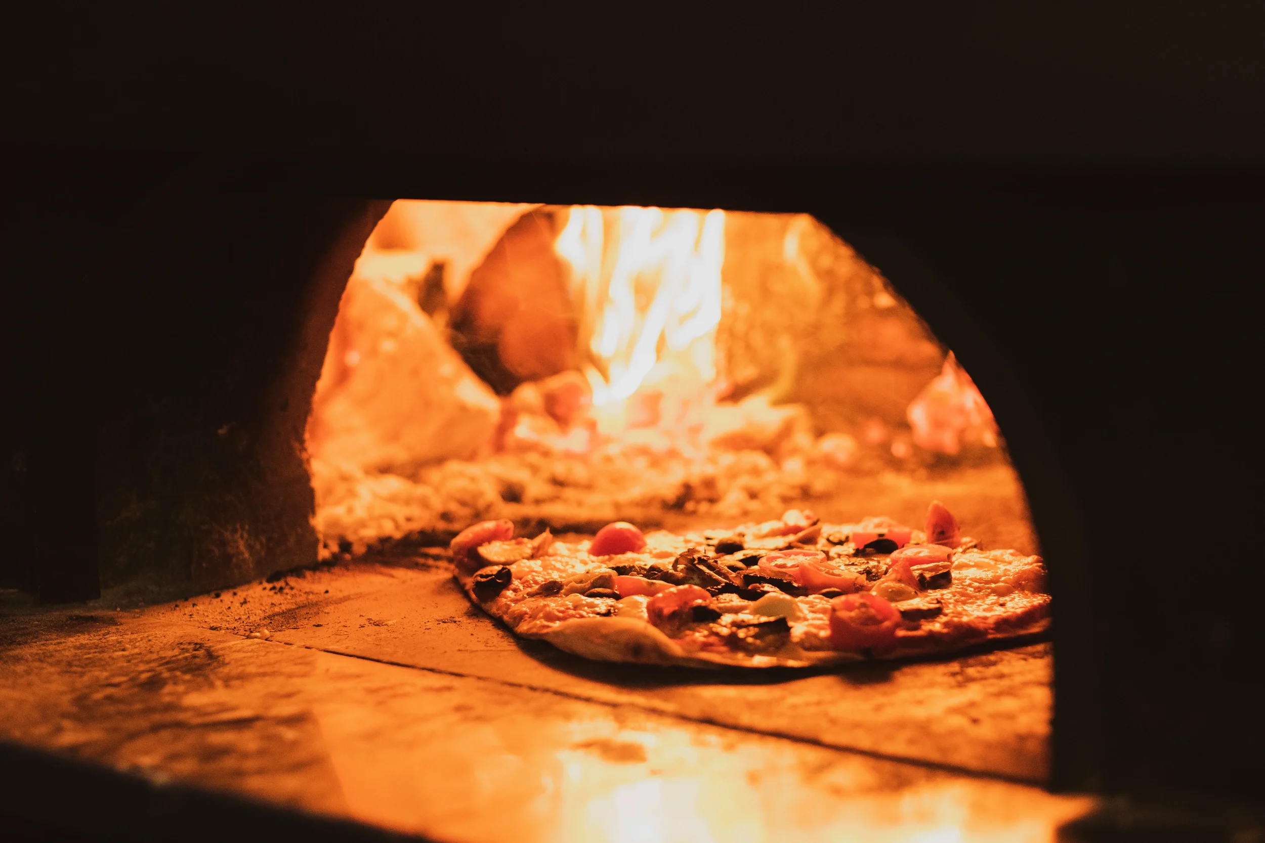 Pizza topped with tomatoes and vegetables baking on the stone floor of a wood-fired oven