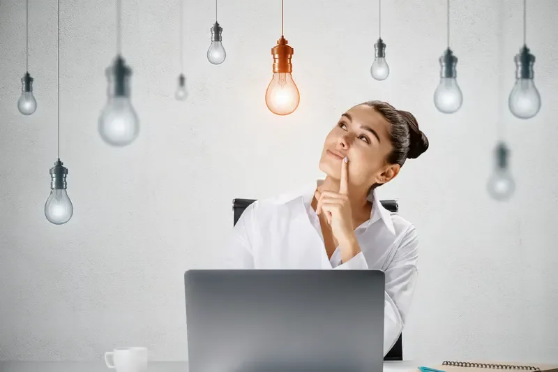 Thoughtful businesswoman sitting at a desk with a laptop looking up at a glowing lightbulb idea concept.