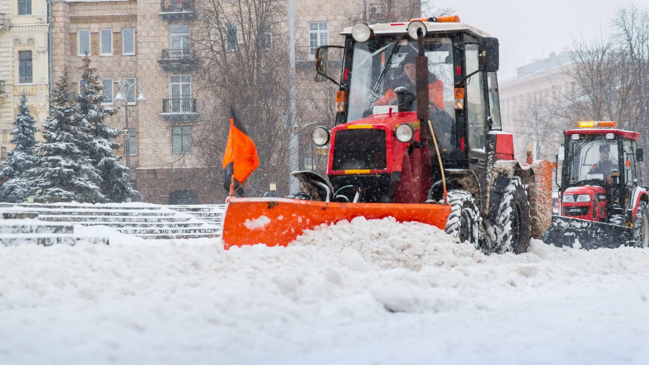 A red industrial tractor equipped with a large orange plow clearing deep snow from a city street during a winter storm.