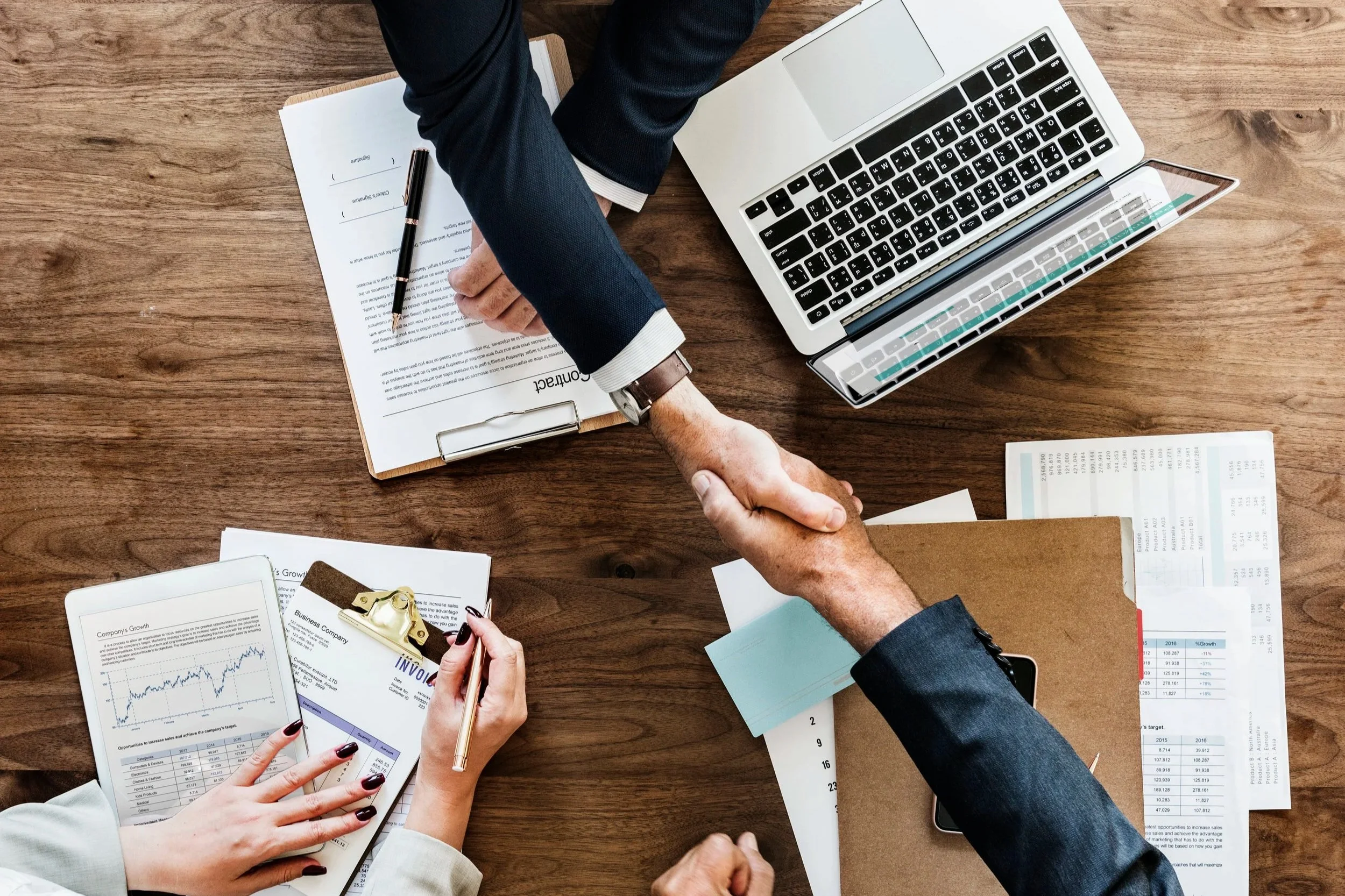Top-down view of professionals shaking hands over a wooden desk with a laptop and signed contracts.