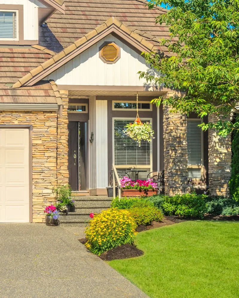 Landscaped front yard and entrance of a stone home featuring vibrant flower beds, green lawn, and potted plants.