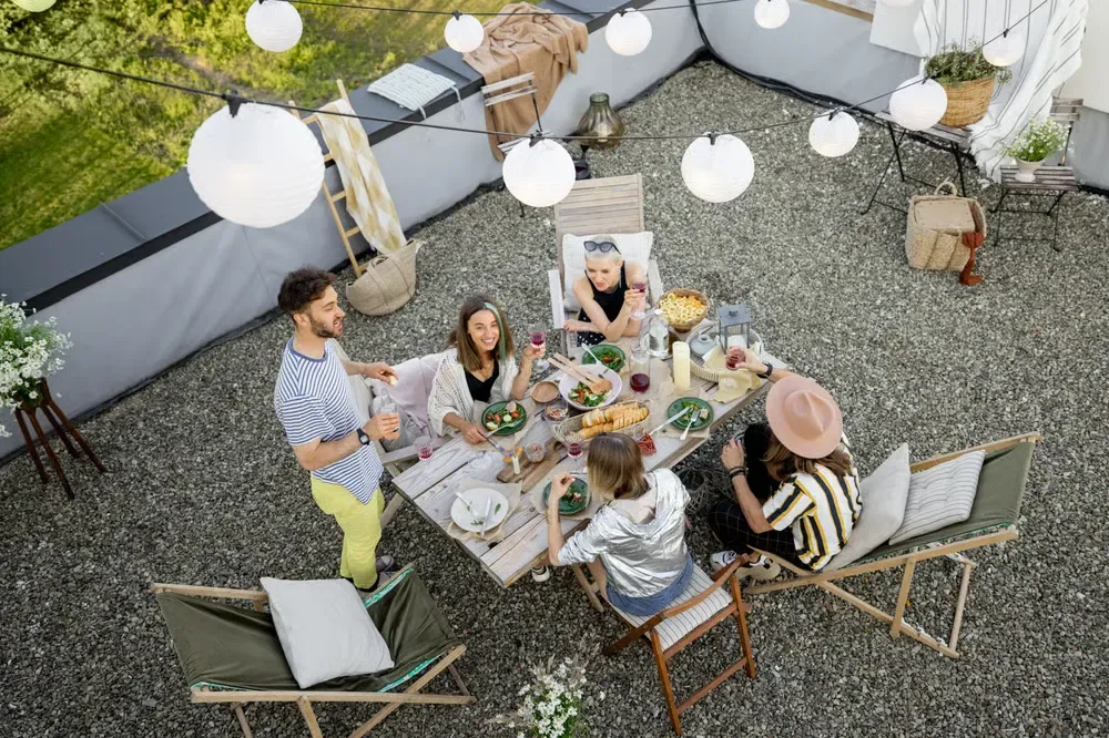 Friends enjoying a dinner party at a wooden table on a gravel rooftop patio under hanging globe lights.