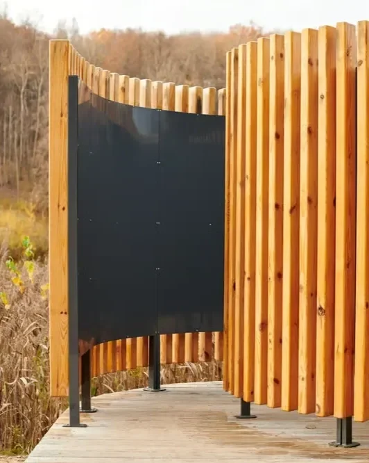 Modern curved privacy fence featuring vertical wooden slats and a dark metal panel accent along an outdoor boardwalk.