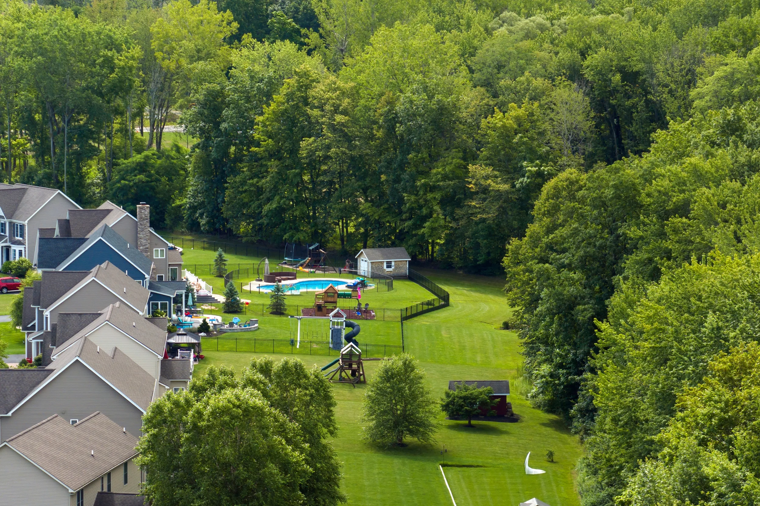 Gemini said Aerial view of a suburban neighborhood with houses bordering a large grassy field with a swimming pool and playground, next to a dense forest.