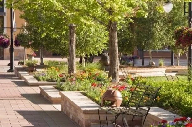A sunlit plaza featuring stone planters with colorful flowers, leafy trees, and black metal benches along a brick walkway.