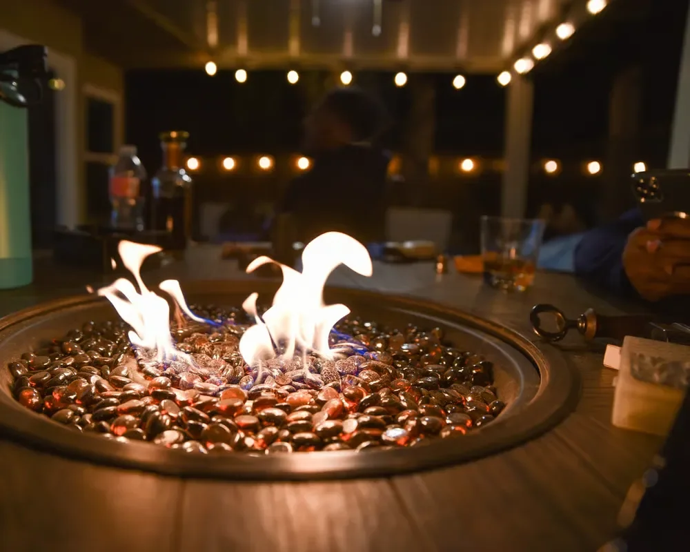 Gas fire pit table with glowing flames and glass beads lit up at night under string lights on a patio.