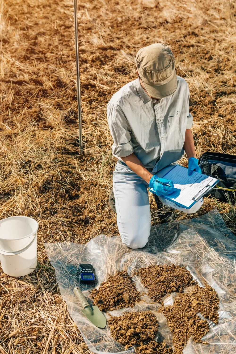 Lawn specialist kneeling in a dry field writing on a clipboard during a professional soil sampling test.