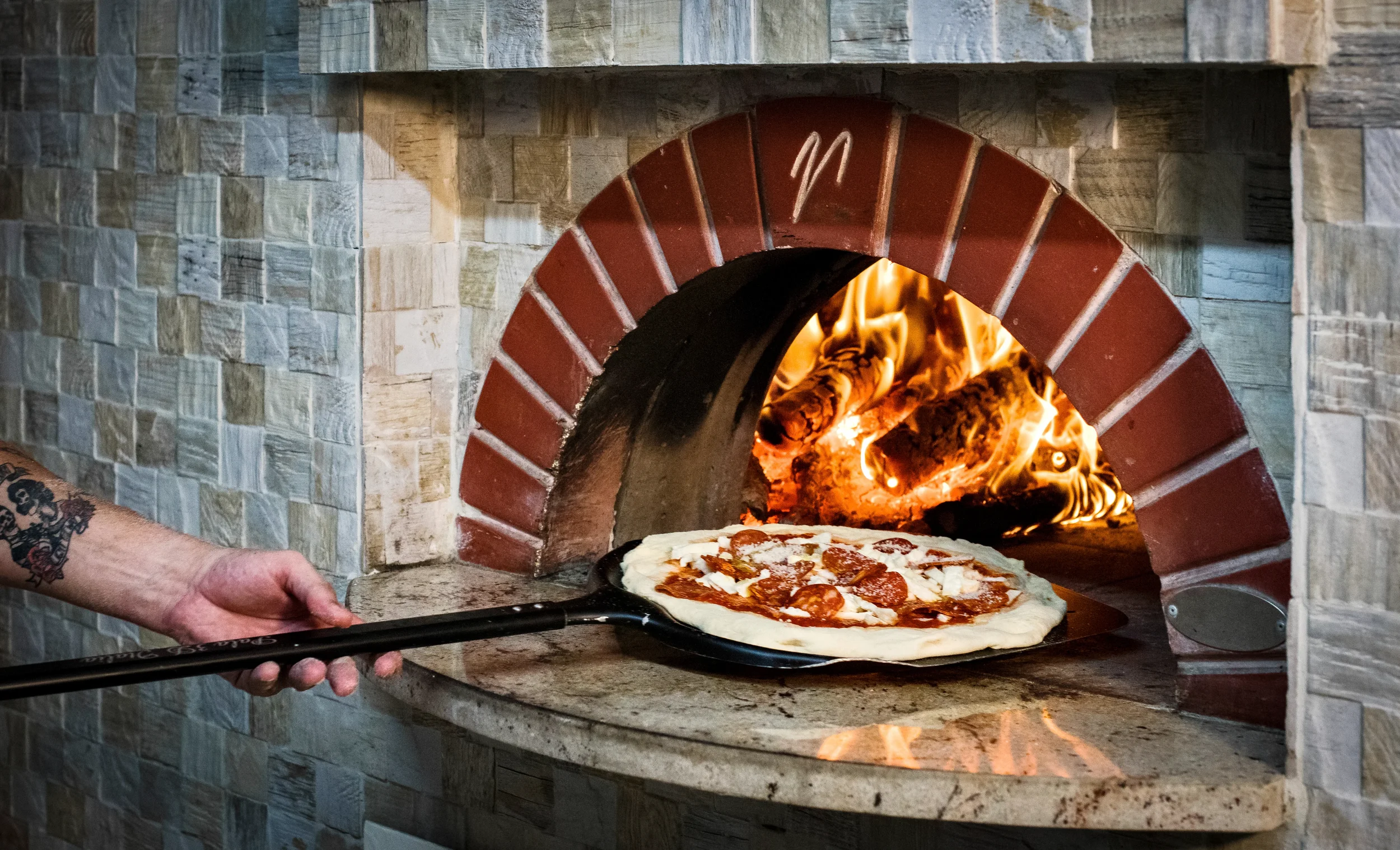 Pepperoni pizza on a peel going into a wood-fired oven with a red brick arch and stone tile wall