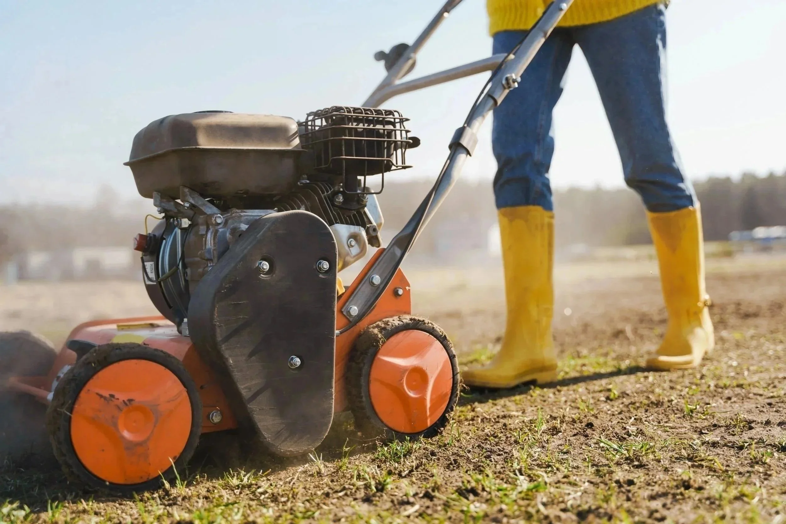 Person in yellow boots operating an orange power tiller to cultivate and prepare soil for planting in a garden.