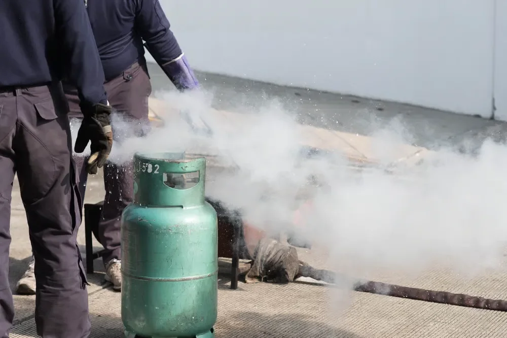 Personnel in safety gear standing near a green propane gas cylinder releasing a plume of vapor during a safety drill.