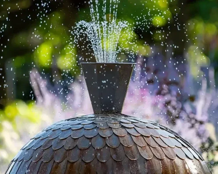 Close-up of a scalloped dome garden water fountain spraying water outdoors with blurred foliage in the background.
