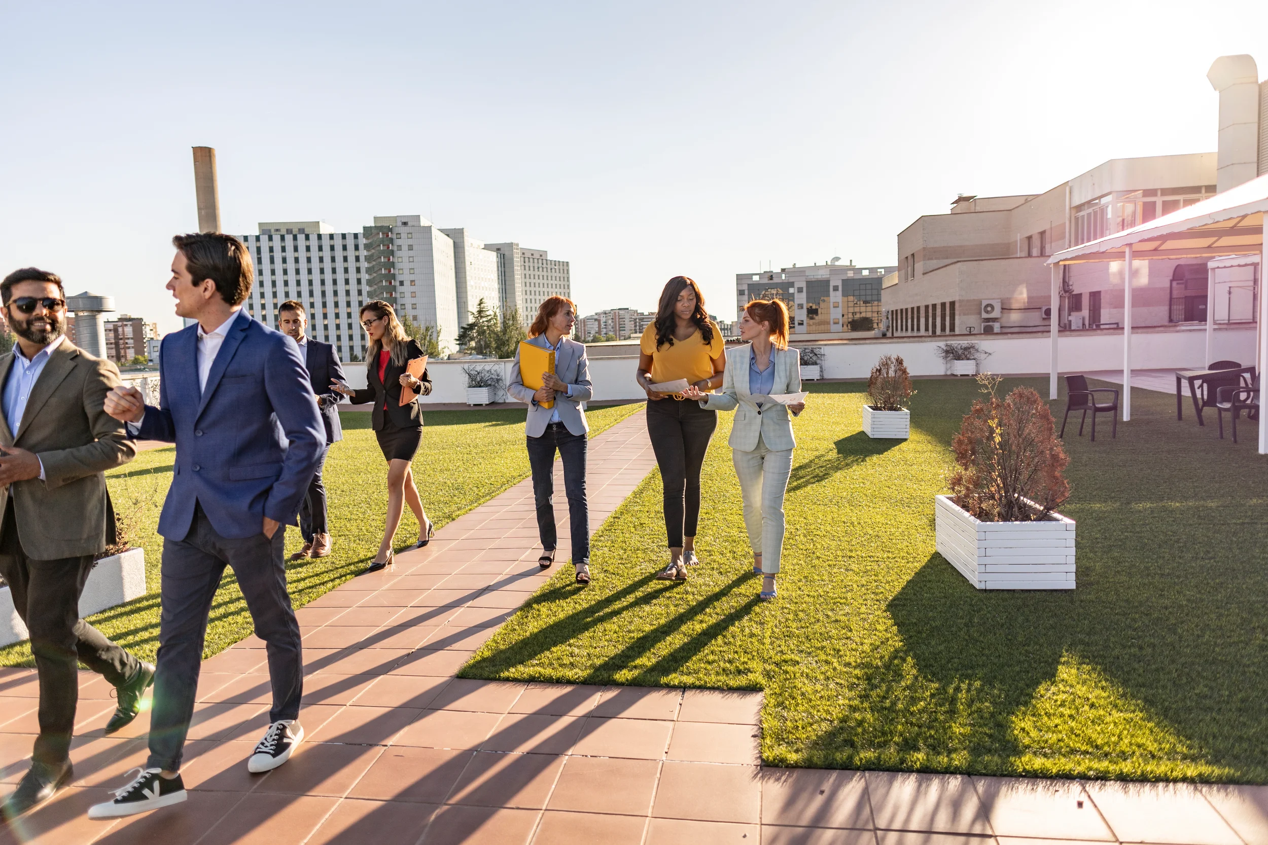 A diverse group of professionals walking and talking across a landscaped courtyard with green turf, surrounded by city buildings.
